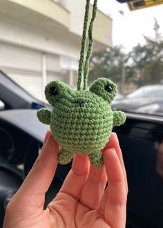 a hand holding a small crocheted frog ornament in front of a car