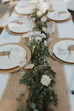 a long table is set with place settings and flowers on the runner, along with silverware