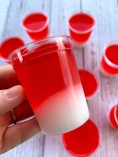 a hand holding a red and white drink in front of small cups on a table