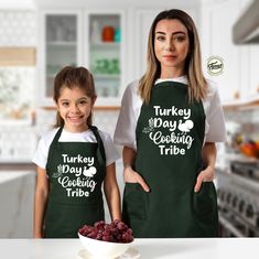 two women wearing green aprons standing next to each other in front of a bowl of grapes