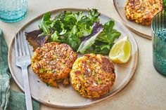 two crab cakes are on a plate with greens and lemon wedges next to it