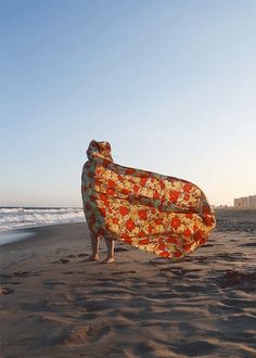 a person standing on top of a sandy beach holding a blanket over their head in front of the ocean