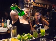 two women sitting at a bar, one drinking from a bottle and the other eating