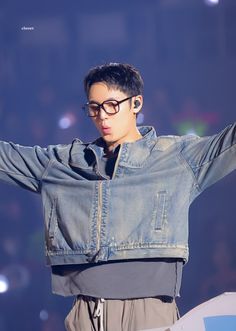 a young man wearing glasses and a denim jacket on stage with his arms stretched out