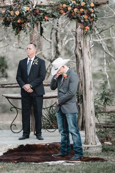 two men are standing under an arch with flowers on it and one man is wearing a cowboy hat