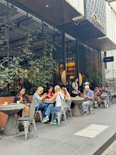 many people are sitting at tables outside in front of a building with glass doors and windows