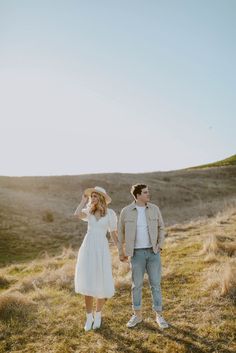 a man and woman holding hands while standing on top of a grass covered hill in the sun