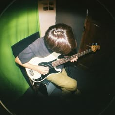 a young man playing an electric guitar in front of a green curtained room with his head down