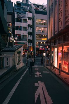 two people walking down the street in front of some tall buildings at dusk with lights on