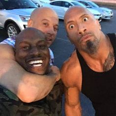 three men are posing for a selfie in front of a parked car and one man has his arm around the other guy's shoulder