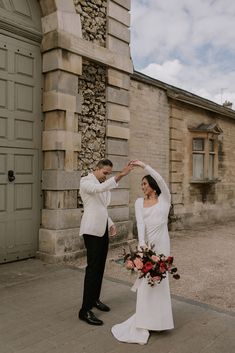 a bride and groom standing in front of an old stone building holding their hands together