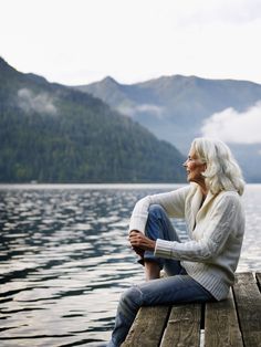 a woman sitting on a dock next to the water with mountains in the back ground