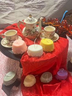 a table topped with tea cups and cakes covered in red cloths next to candles