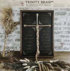 a wooden cross sitting on top of a table next to dried flowers and feathers in front of a sign that reads trinity braid