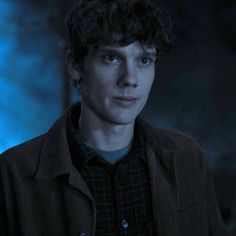 a young man with curly hair and blue eyes looks at the camera in a dark room