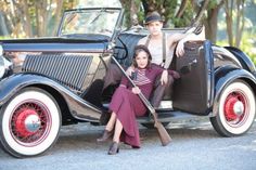 a man and woman sitting on the back of an old fashioned car, posing for a photo