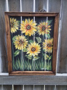 a painting of yellow sunflowers on a black background in a wooden frame hanging on a fence