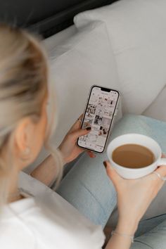 a woman sitting on a couch holding a cup of coffee and looking at her cell phone