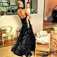 a woman in a black dress holding a baby while standing next to a couch and chair