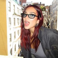 a woman with red hair and sunglasses on looking at something in the distance while standing next to a building