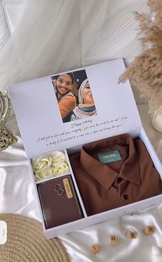 an open gift box containing a brown shirt and some other items on a white bed