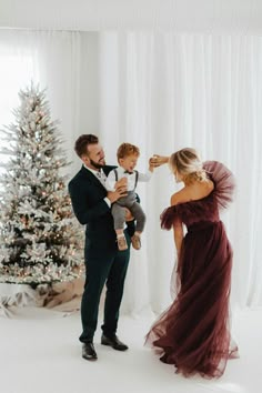 a man in a tuxedo holds his son as he stands next to a christmas tree