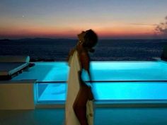 a woman in a white dress standing next to a swimming pool at night with the sun setting