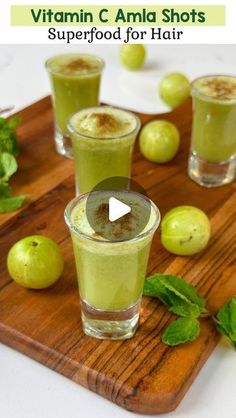 two glasses filled with green smoothie on top of a wooden cutting board next to fruit