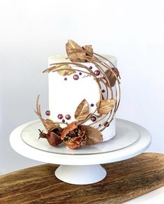 a white cake with gold leaves and berries on top sitting on a wooden platter