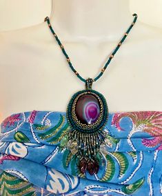 When I ran across this cabochon I had to have it. I crafted this necklace in bead embroidery with deep teal beads to compliment the intense purple color of the cabochon. I added sweet pressed glass leaves on the fringe. This is a one of a kind piece and if you are a purple lover, like me, it will not disappoint! The pendant from loops to fringe measures 4 inches long and 2 inches wide. The seed bead necklace is 21 inches long and features a gold tone lobster claw clasp. Bohemian Oval Beaded Necklaces, Bohemian Oval Beaded Necklace, Handmade Oval Beaded Bohemian Necklaces, Handmade Bohemian Oval Beaded Necklaces, Handmade Purple Pendant Beaded Necklace, Bohemian Beaded Necklaces With Cabochon For Gifts, Handmade Purple Beaded Pendant Necklace, Oval Beaded Gems For Jewelry Making, Embroidery Necklace