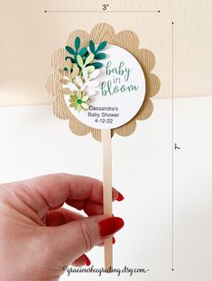 a hand holding a wooden stick with a flower on it that says baby in bloom