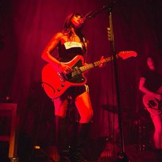 a woman in short shorts and boots playing guitar on stage with other musicians behind her