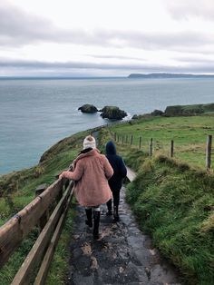 two people walking down a path next to the ocean on top of grass covered hills