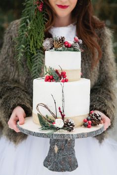 a white wedding cake with pine cones and berries on top is held by two hands