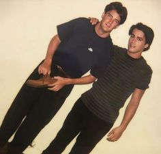 two men standing next to each other in front of a white wall with one man holding his hat