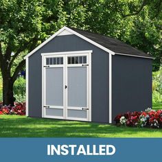 a gray shed with the words installed above it and flowers in front of it on grass