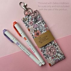 two pens sitting next to each other on top of a pink surface with a keychain