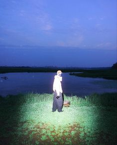 a man standing on top of a lush green field next to a lake at night