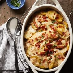 a casserole dish with potatoes and bacon in it on a table next to spoons