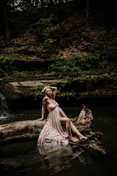 a woman in a dress sitting on a log in the water near a small waterfall