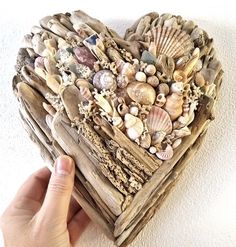 a heart shaped box filled with seashells on top of a white wall next to a person's hand