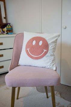 a pink chair with a smiley face pillow on it's back in front of a white dresser