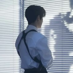 a man standing in front of a window with his back turned to the camera, wearing a white shirt and black suspenders