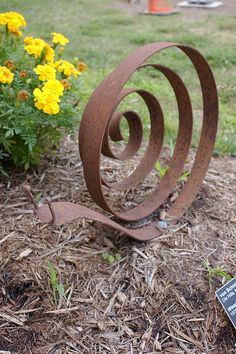 a rusted metal sculpture sitting in the middle of a flower bed next to yellow flowers