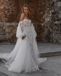 a woman wearing a white wedding dress with sheer sleeves and flowers on the skirt is standing in front of a stone wall