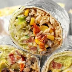 three burritos wrapped in aluminum foil on top of a wooden table with rice and vegetables