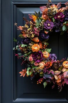 a wreath with purple and orange flowers is hanging on a black front door, surrounded by greenery