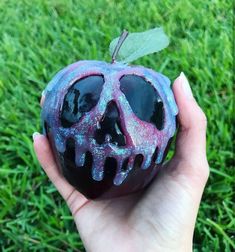 a hand holding an apple shaped like a skull on top of green grass with blue and pink glaze