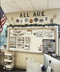 an all are welcome sign on the wall above a computer desk in front of a flag