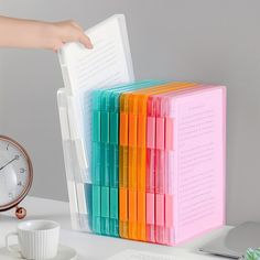 a stack of colorful file folders sitting on top of a desk next to a clock